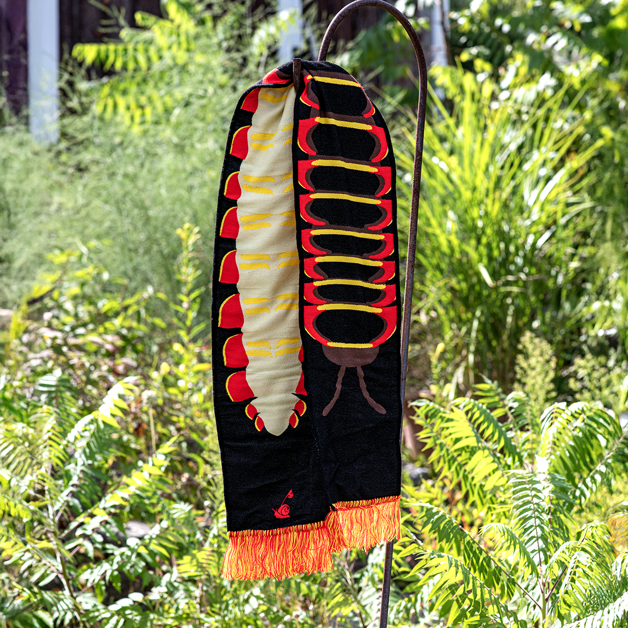 Cherry Millipede scarf hanging on a hook against a green foliage background
