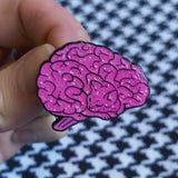 A soft enamel pin in the shape of a human brain. Upon closer inspection, the lines of the brain resemble a sleeping cat. A woman's fingers move the pin, showing it glitter and glisten. In the background is a houndstooth print.