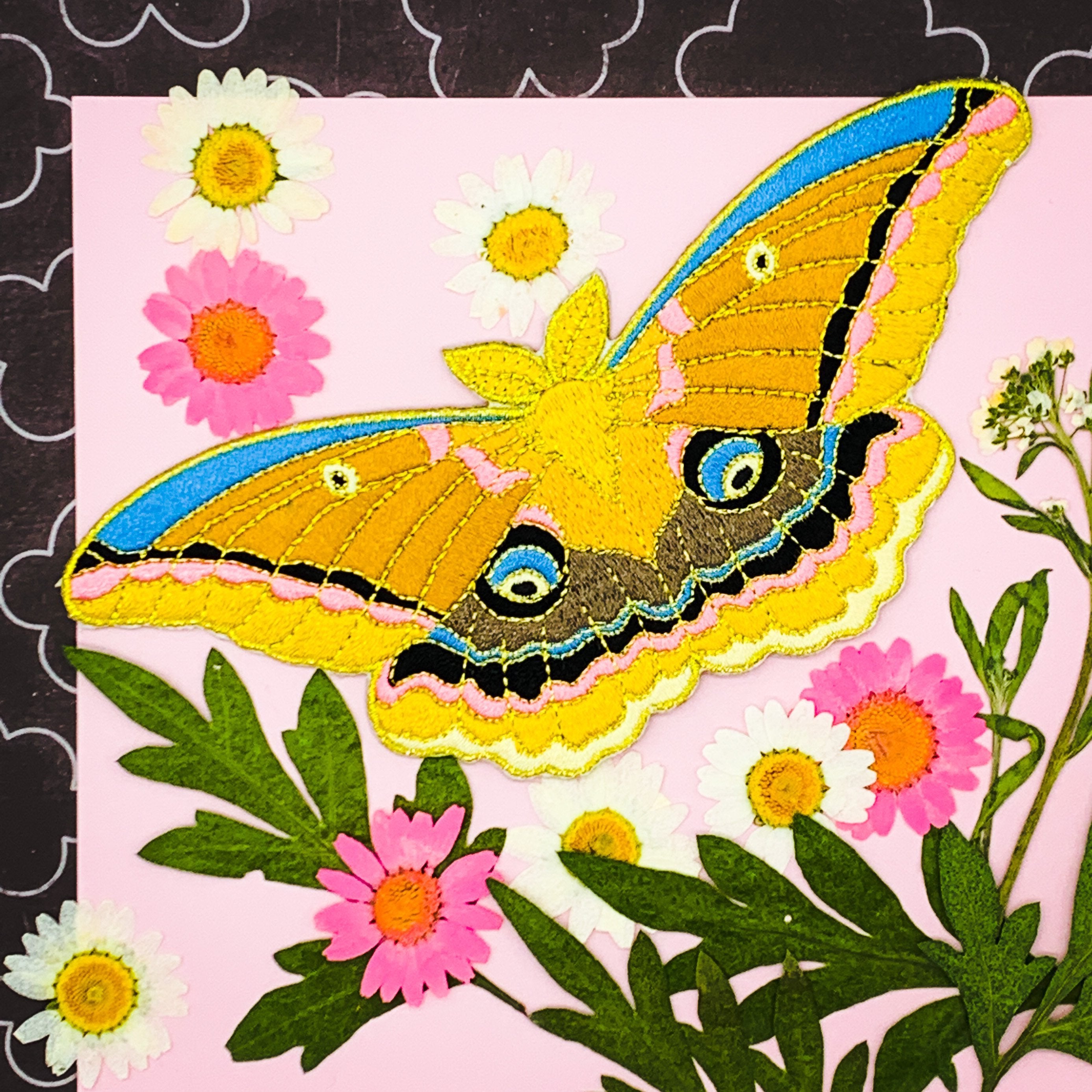 A soft embroidered patch of a polyphemus moth, laid out on decorative paper with colorful dried flowers.