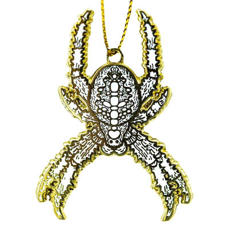 A gold and white enamel holiday tree ornament of a cross orb weaver Christmas Spider danging from a golden string on a white background.