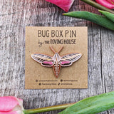 An enamel pin of a brown, bronze, and pink white lined sphinx moth, with wings outstretched.