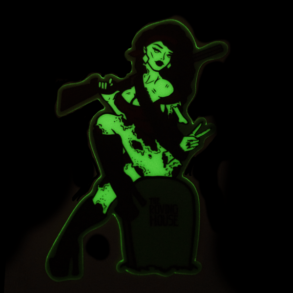 A vinyl sticker of an attractive, buxom zombie woman sitting on a gravestone and holding a shotgun. The sticker glows green in the dark.