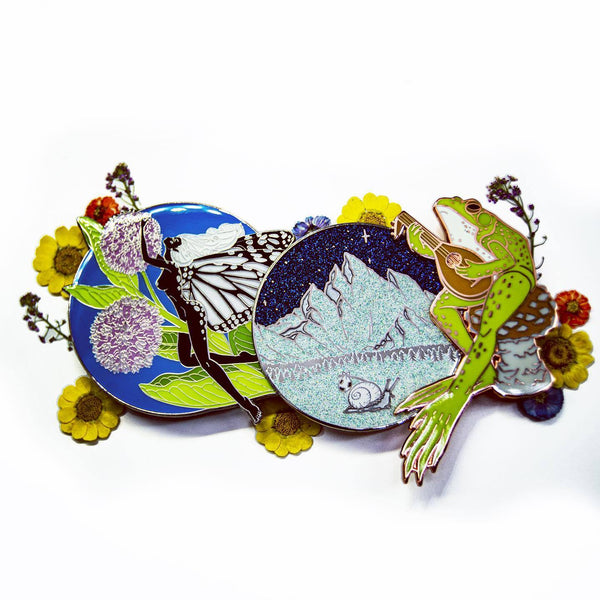 Three very large enamel pins surrounded by dried flowers.