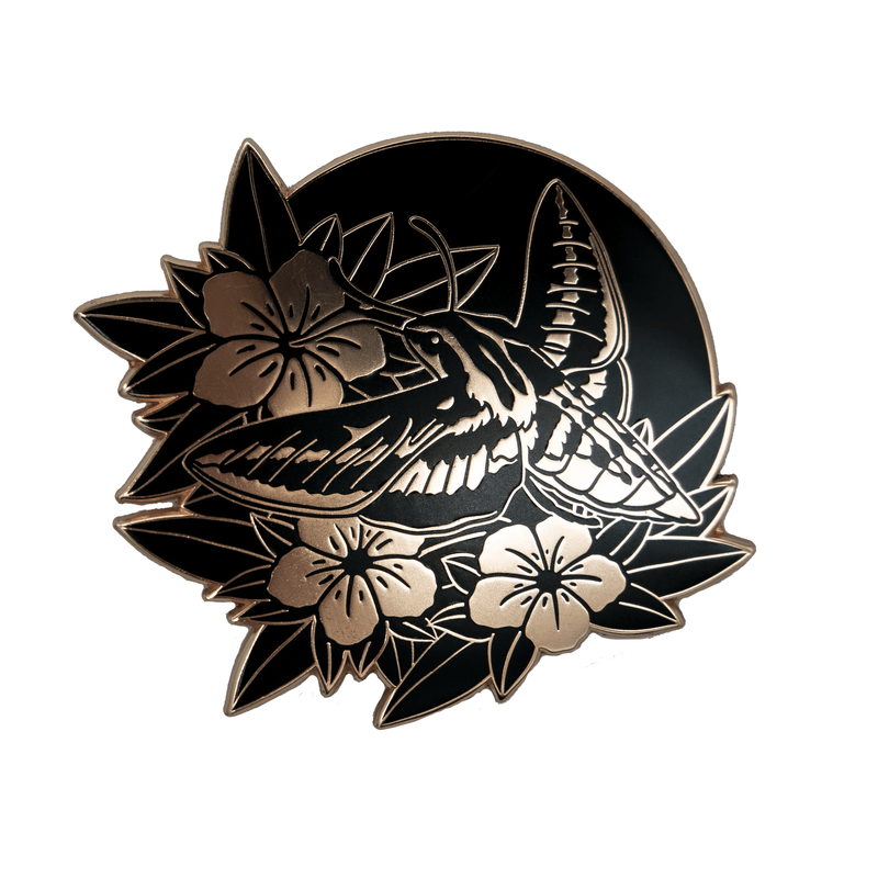 A large, rose gold and black enamel pin of a white lined sphinx moth hovering like a hummingbird over wild petunias.