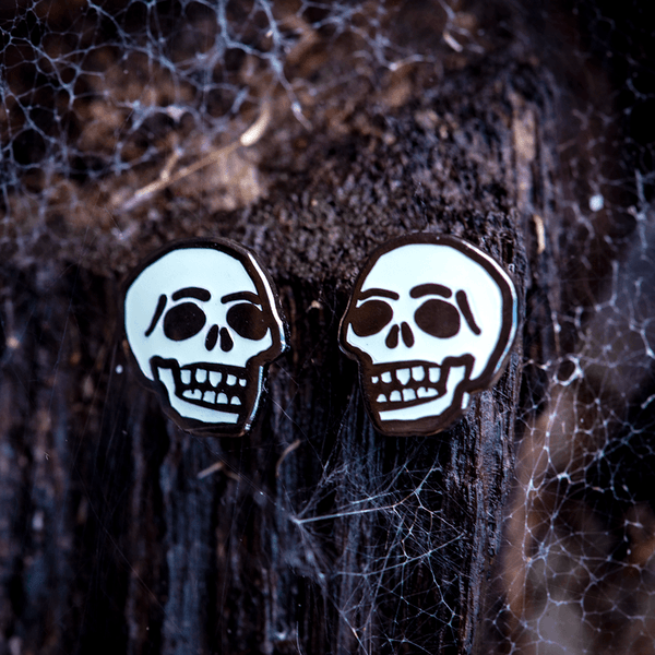 Little Skulls Pin Set by The Roving House. This mirrored pair of hard enamel pins features stylized white skulls on black nickel metal. The pins glow in the dark.