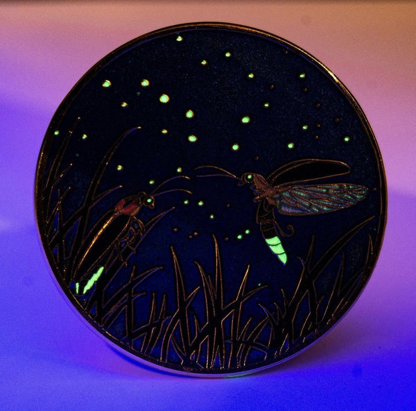 An dim-light view of an enamel pin. Two Big Dipper fireflies meet under the night sky, with their abdomens and the constellations above glowing green..