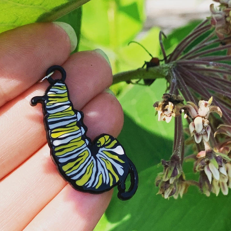 An enamel charm of a black, white, and yellow monarch caterpillar hanging in the J shape. Behind it is a milkweed plant, with a milkweed bug watching.
