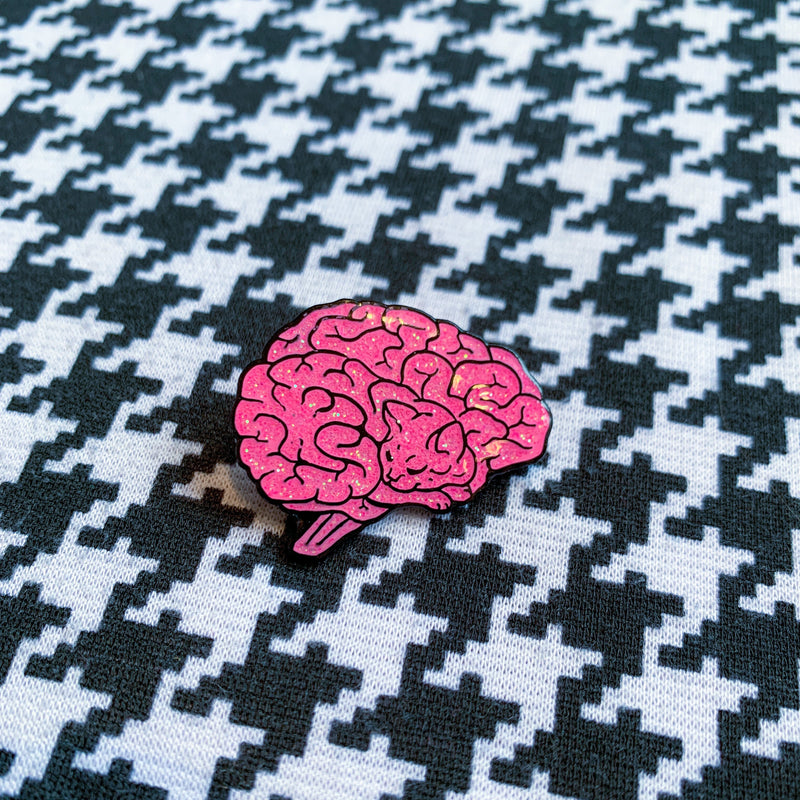 A soft enamel pin in the shape of a human brain. Upon closer inspection, the lines of the brain resemble a sleeping cat. The pin sparkles with glitter and glistens with an epoxy coating. In the background is a houndstooth print.