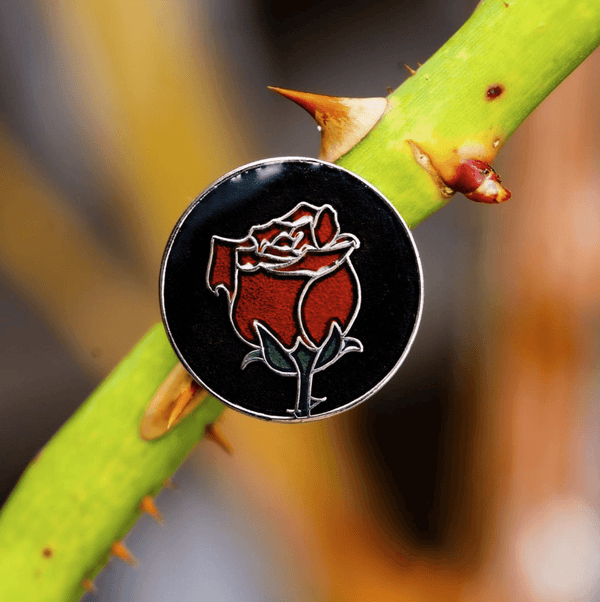 A round pin with black background and a red rose that resembles stained glass art. The pin is attached to a real rosebush branch.