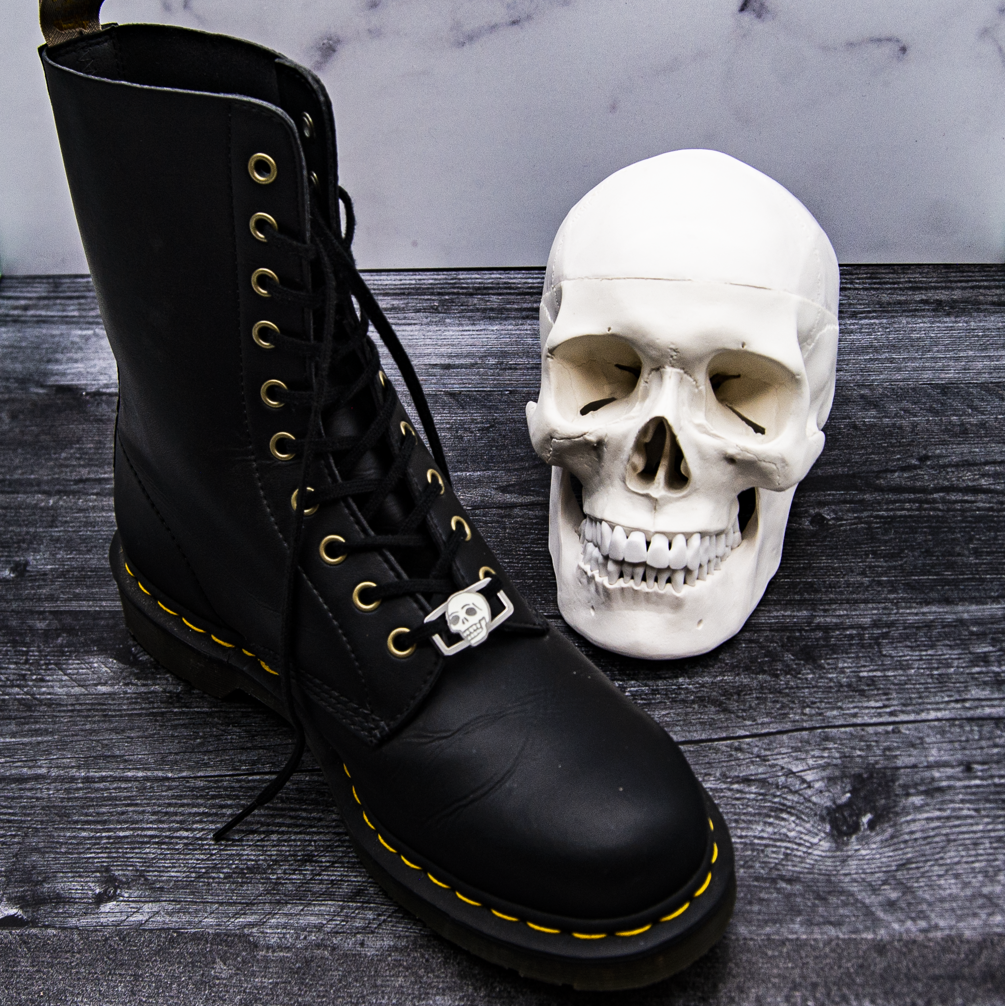 A classic black Doc Marten boot displaying a skull shoelace charm neck to a model of a human skull.