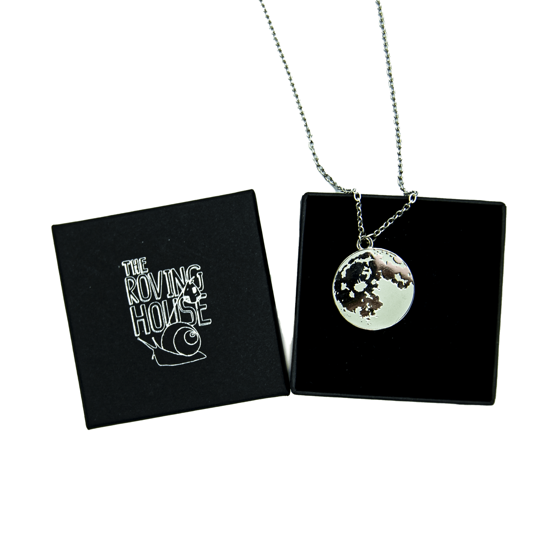 A grey enamel and black nickel pendant of the full moon on a silver metal chain, draped over a black jewelry box with The Roving House snail logo printed on its cover.