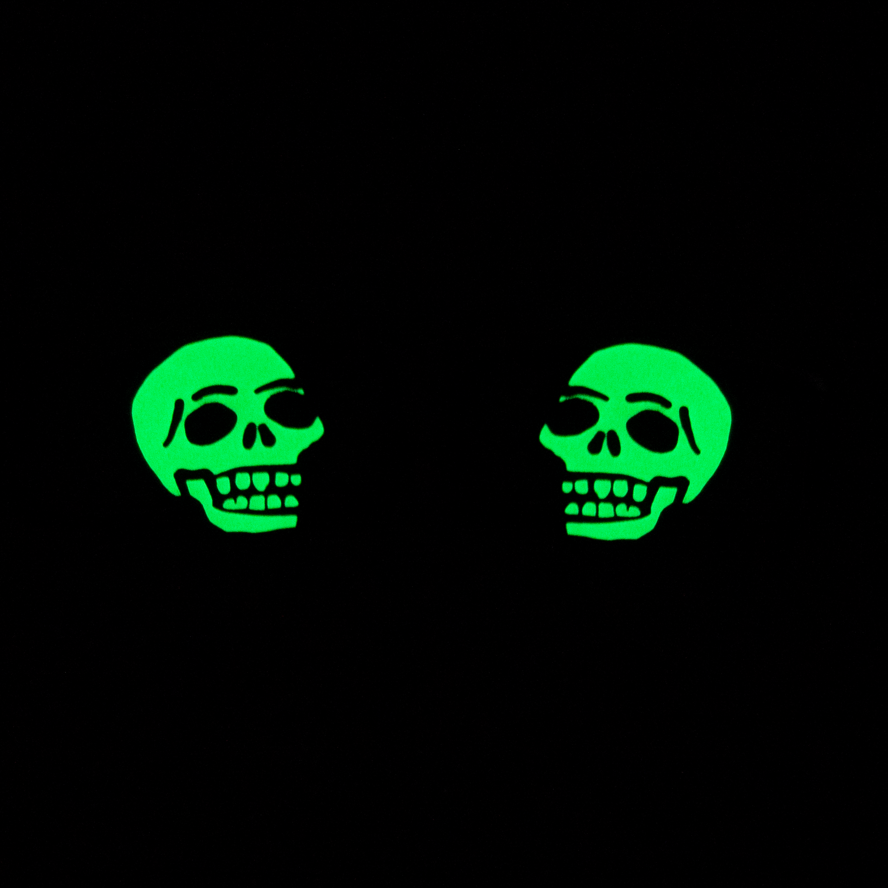 A pair of mirrored skull shoelace charms glowing green in the dark.
