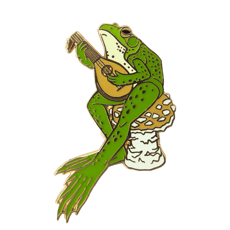 An enamel pin of a northern green frog sitting atop a golden agaric mushroom, strumming a medieval stringed instrument.