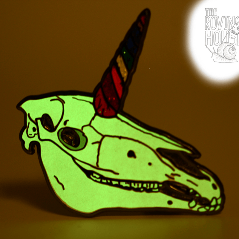 A hard enamel pin of unicorn skull with a rainbow swirled horn. The skull is glowing in dimmed light.