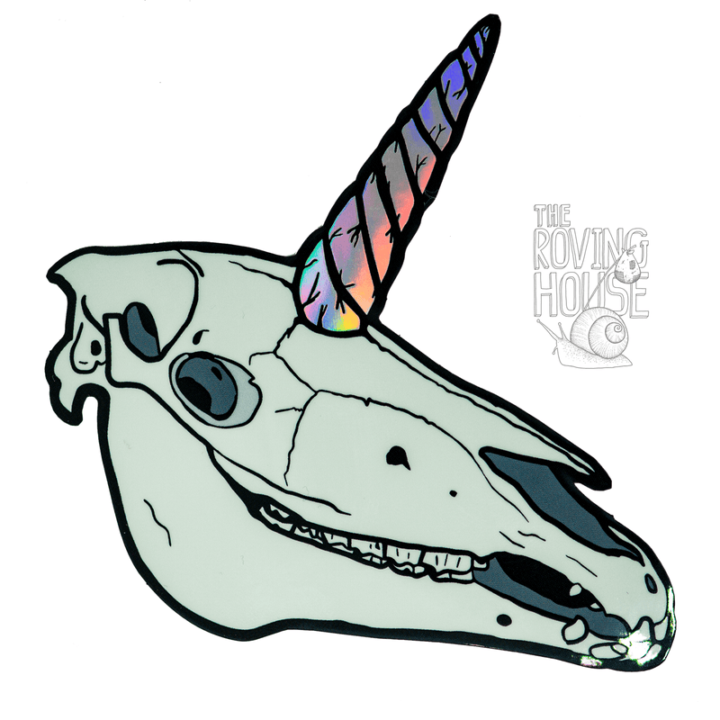 A vinyl sticker of a unicorn skull with a holographic rainbow swirled horn. The skull is glowing in dimmed light.