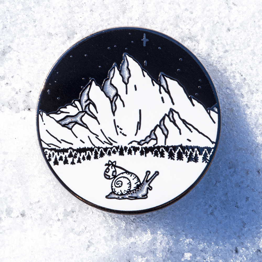 Enamel pin featuring The Roving House snail with his bindle or hobo bag, exploring snowy mountains beneath the northern stars.